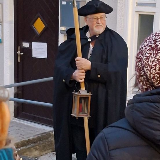 Ein älterer Mann in einem schwarzen Gewand und Hut hält draußen eine Laterne in der Hand.