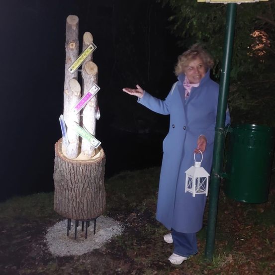 Eine ältere Frau in einem blauen Mantel hält eine Laterne in der Hand, während sie neben einer Holzstruktur steht. An der Struktur sind mehrere Schilder angebracht. Ein grüner Mülleimer steht rechts.