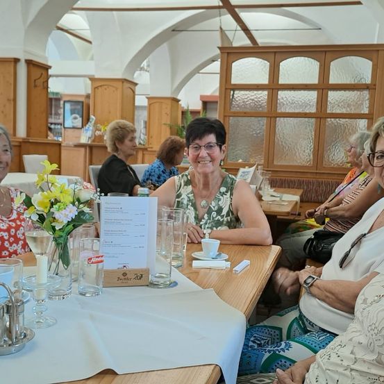 Eine Gruppe von Frauen sitzt an einem Tisch in einem Restaurant. Sie lächeln und genießen ihr Essen. Auf dem Tisch steht eine Blumenvase, zusammen mit einer Speisekarte.