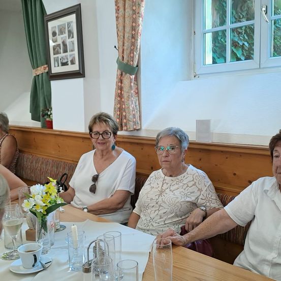 Drei ältere Frauen sitzen an einem Tisch mit einem Blumen-Centerpiece. Sie lächeln und scheinen ihre Mahlzeit zu genießen. Der Raum hat Holzwände, Vorhänge und ein gerahmtes Bild an der Wand.