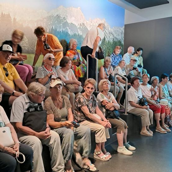 Eine Gruppe älterer Frauen sitzt in einer Reihe, einige schauen auf ihre Telefone. Eine Frau steht in der Nähe des Geländers, und es gibt ein Wandbild eines Berges im Hintergrund.