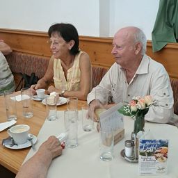 Ein älteres Paar sitzt an einem Tisch, lächelt und schaut sich an, mit Getränken und einer Blumenarrangement vor ihnen.