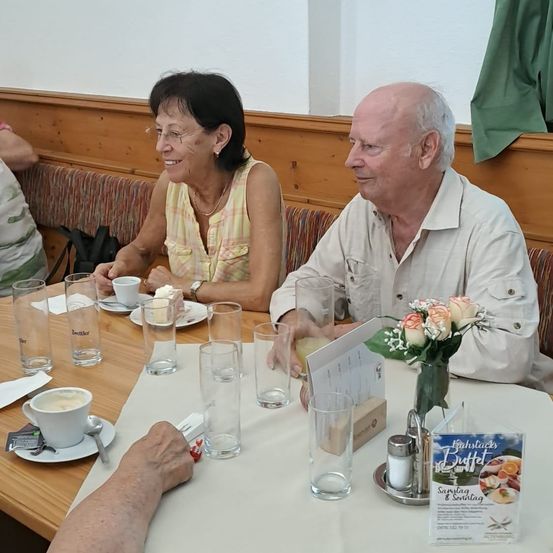 Ein älteres Paar sitzt an einem Tisch, lächelt und schaut sich an, mit Getränken und einer Blumenarrangement vor ihnen.