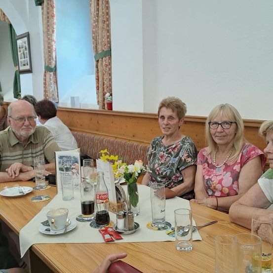 Eine Gruppe von Menschen, darunter Frauen und Männer, sitzt an einem Tisch in einem Restaurant. Der Tisch ist mit Tellern, Gläsern und Blumen gedeckt. Die Frau in der Mitte trägt eine Brille und eine Halskette. Hinter ihnen befindet sich ein Fenster mit einer Vase auf der Fensterbank.