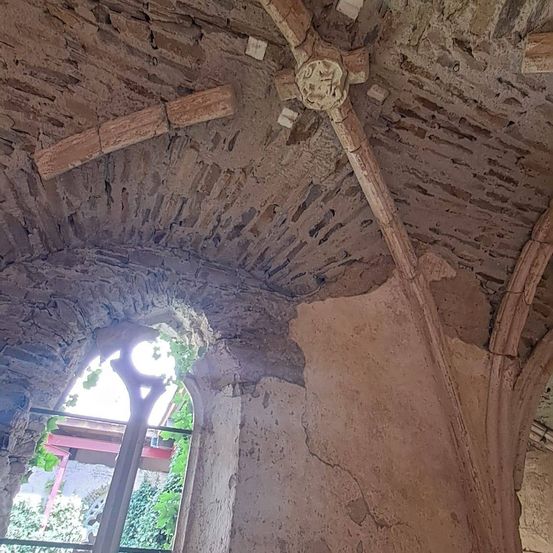 Eine alte architektonische Struktur mit verwitterten Steinmauern und Holzbalken. Die Decke zeigt Anzeichen von Alter und Beschädigung. Ein Fenster mit einem Bogen offenbart einen Blick nach draußen.
