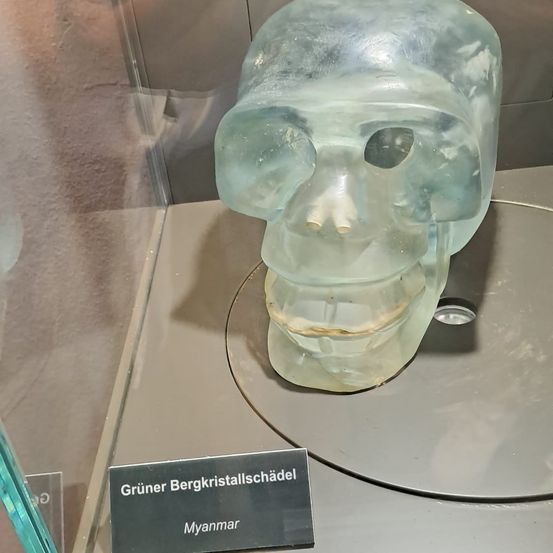 Ein grüner Kristallschädel, möglicherweise aus Myanmar, ist in einem Museum ausgestellt. Der Schädel ist durchsichtig mit sichtbaren Zähnen und einem Lächeln. Er sitzt auf einer runden Platte.