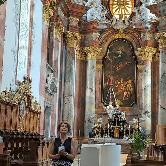 Eine Frau steht in einer großen Kathedrale und betrachtet ein kunstvolles Altarbild mit goldenen Schnitzereien, Statuen und einem Buntglasfenster darüber.