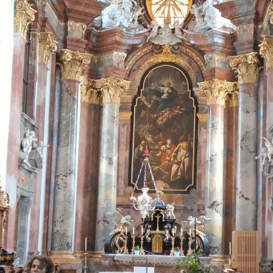 Ein großes Altarbild dominiert ein barockes Kircheninneres mit verzierten Säulen und Marmorwänden. Ein komplizierter Kronleuchter hängt über einem detaillierten Gemälde. Statuen sind an den Wänden angebracht.