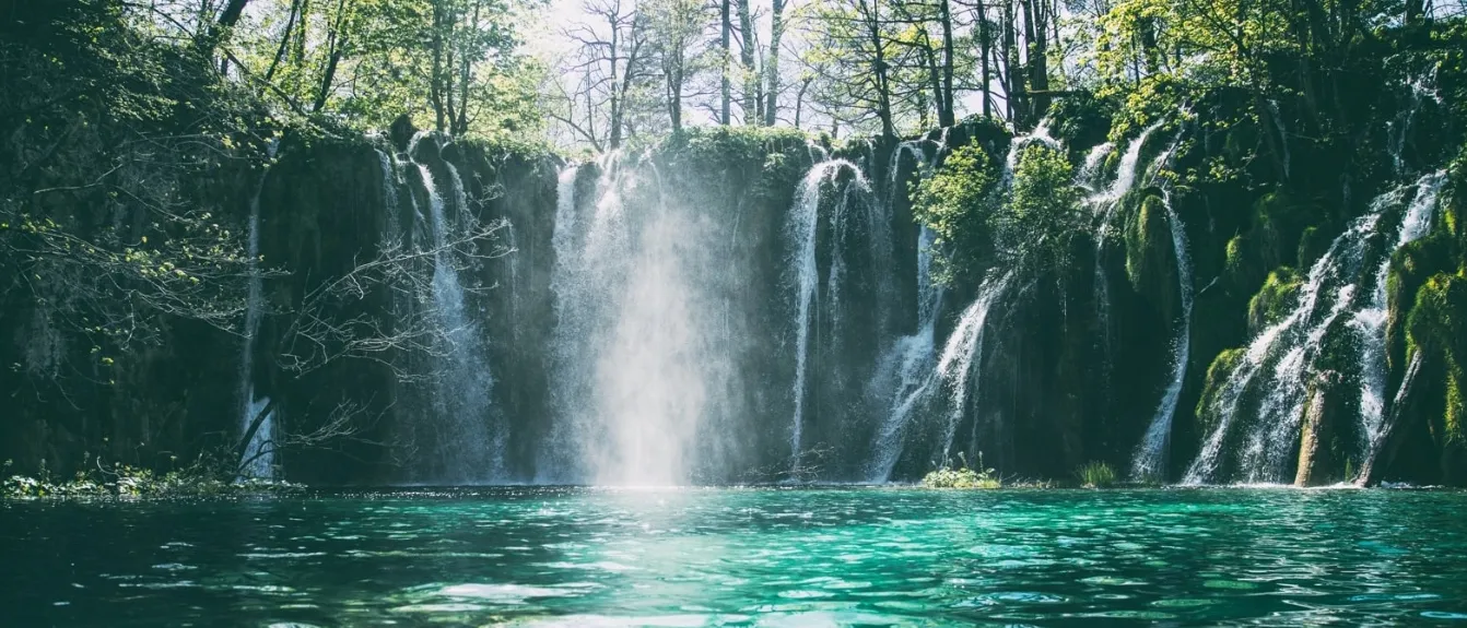 Ein atemberaubender Wasserfall stürzt von einem felsigen Abhang in klares blaues Wasser, umgeben von üppigem Grün unter einem strahlenden Himmel.