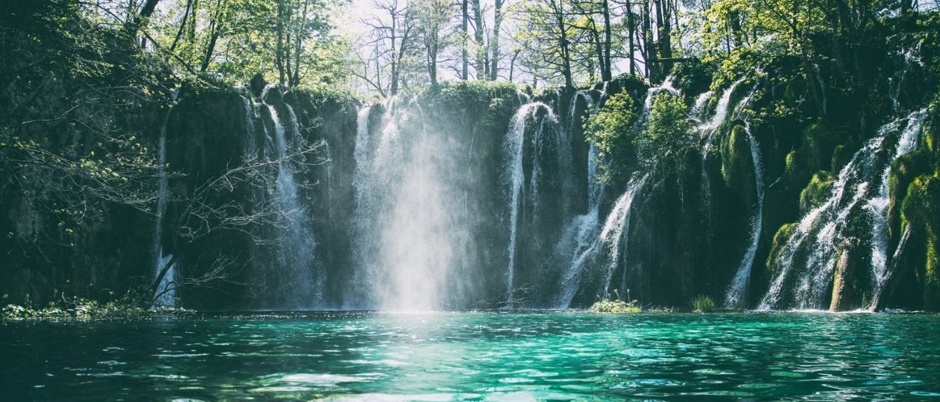 Ein atemberaubender Wasserfall stürzt von einem felsigen Abhang in klares blaues Wasser, umgeben von üppigem Grün unter einem strahlenden Himmel.