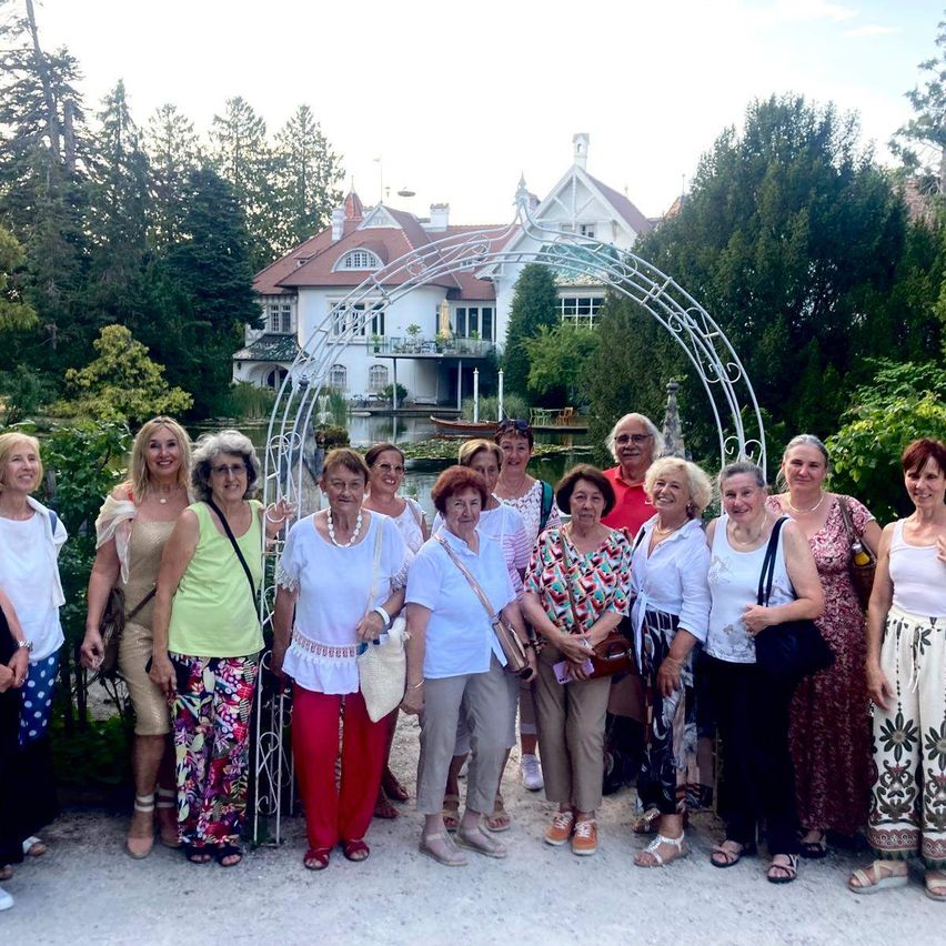 Eine Gruppe älterer Frauen und Männer steht vor einem weißen Haus mit Garten und Brunnen. Sie lächeln und posieren für ein Foto.