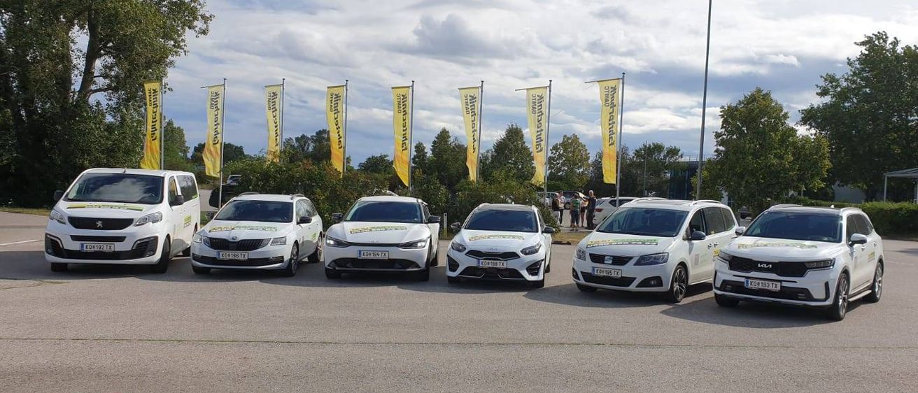 Fünf weiße Autos sind in einer Reihe auf einer Betonfläche geparkt, mit Bannern im Hintergrund. Bäume und Straßenlaternen sind in der Ferne sichtbar.