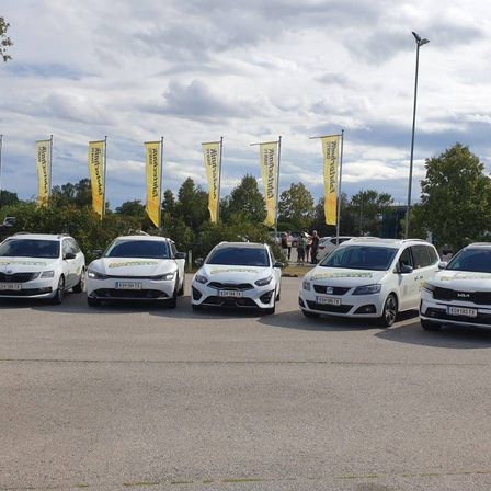 Sechs weiße Autos stehen in einem Parkplatz mit Bannern und Bäumen im Hintergrund. Ein bewölkter Himmel ist sichtbar.