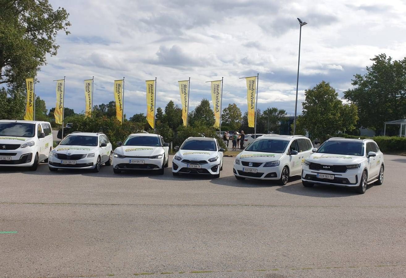 Sechs weiße Autos stehen in einem Parkplatz mit Bannern und Bäumen im Hintergrund. Ein bewölkter Himmel ist sichtbar.