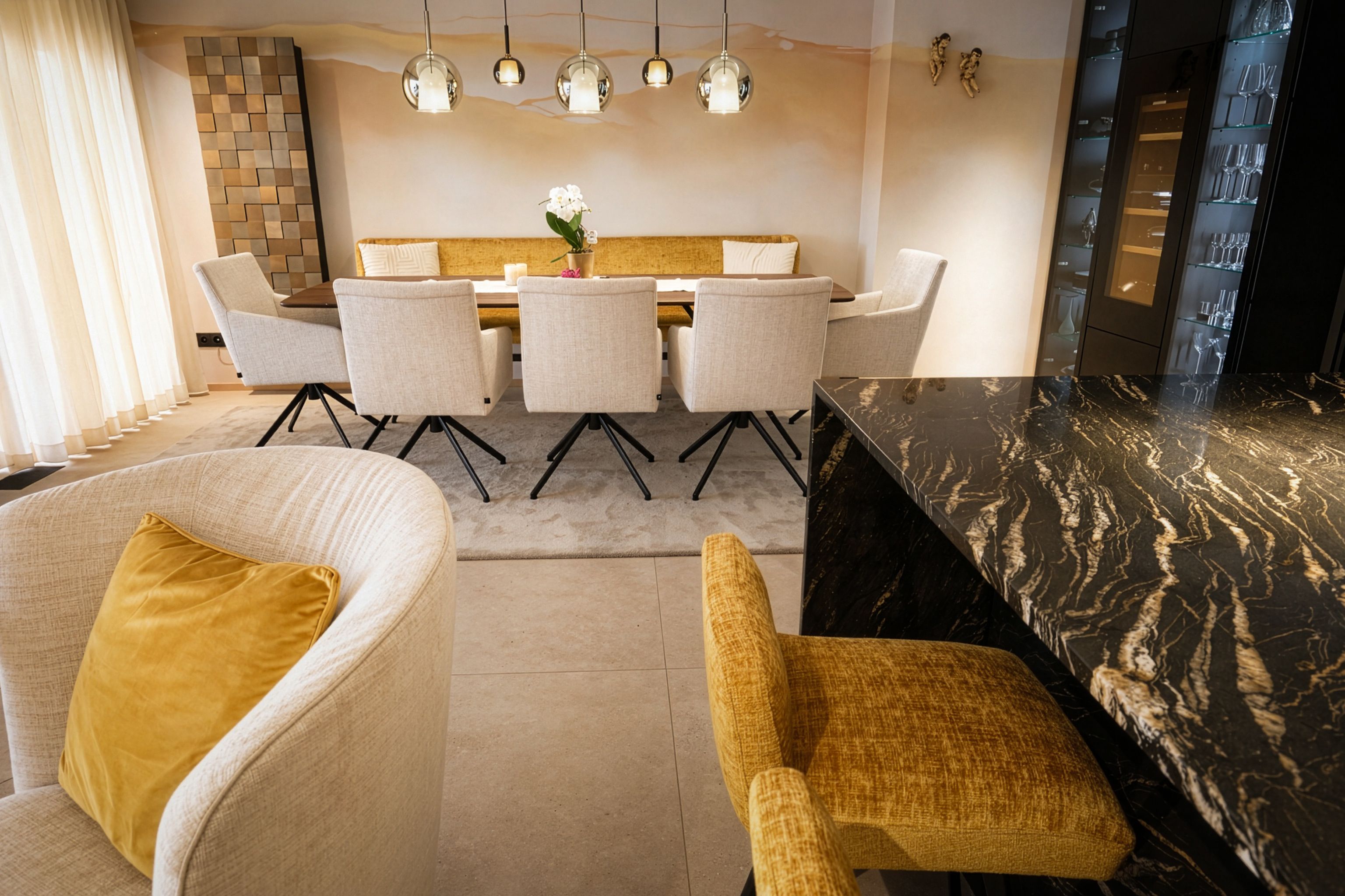 Ein modernes Esszimmer mit einem runden Tisch, umgeben von sechs Stühlen, einer langen Couch, einer Marmortheke und einer beigen Wand mit einem großen Wandbild.