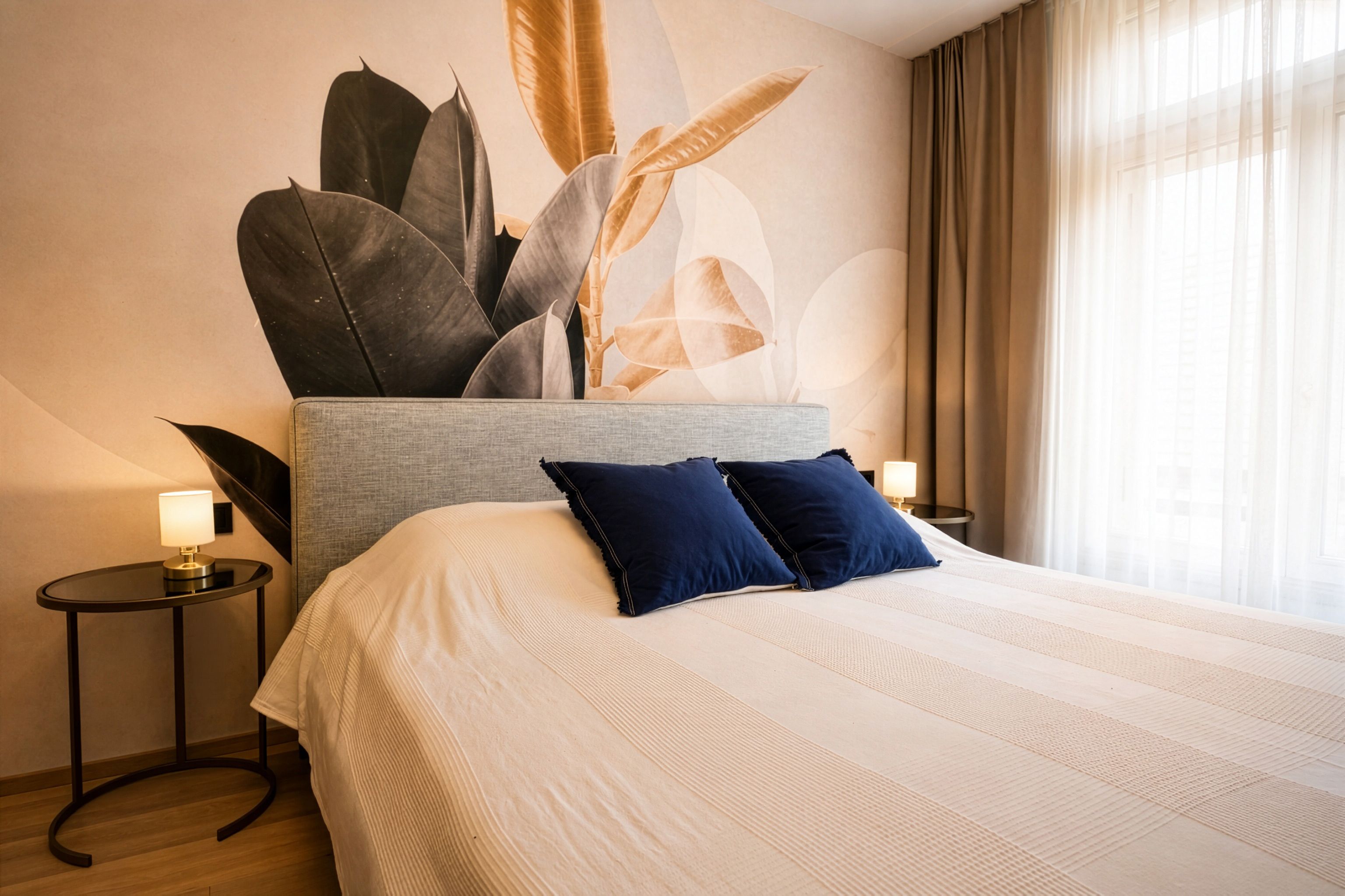 Ein Schlafzimmer mit einem Bett, das mit blauen Kissen verziert ist, vor einer Wand mit einem Tapetenbild von tropischen Blättern.