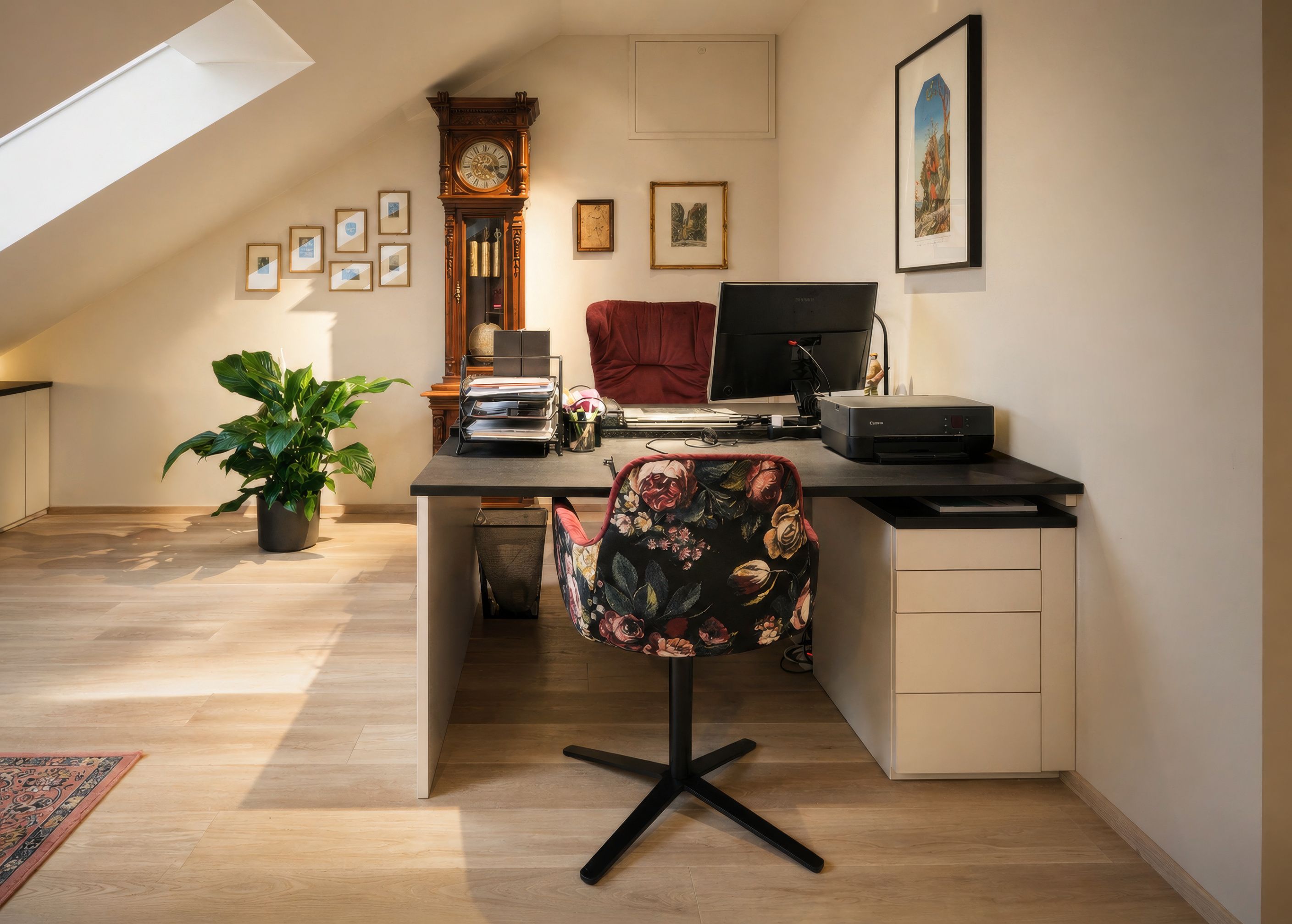 Ein Heimbüro mit schwarzem Schreibtisch, Blumenstuhl und Drucker. Ein Großvater-Uhr, gerahmte Bilder und eine Topfpflanze sind ebenfalls sichtbar.