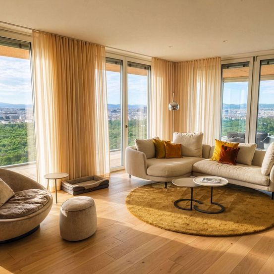 Ein modernes Wohnzimmer mit großen Fenstern, einer beigen Couch mit Kissen, einem runden Teppich und einem Tisch mit Büchern. Eine Lampe und ein Hundebett sind in der Nähe. Die Aussicht zeigt eine Stadtlandschaft.