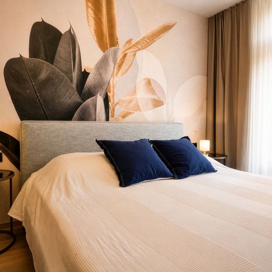 Ein Schlafzimmer mit grauem Kopfteil, weißem Bettlaken und blauen Kissen. Die Wand ist mit einer Tapete aus tropischen Blättern verziert.