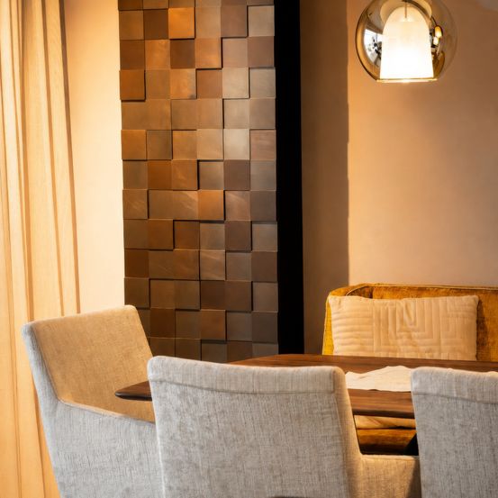 Ein modernes Esszimmer mit brauner gefliester Wand, runder Deckenlampe und gepolsterten Stühlen um einen Holztisch.
