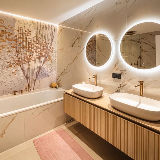 Ein modernes Badezimmer mit Badewanne, zwei Waschbecken und zwei runden Spiegeln mit Beleuchtung. Die Wände sind mit Marmorfliesen und einer dekorativen Wandkunst verziert.