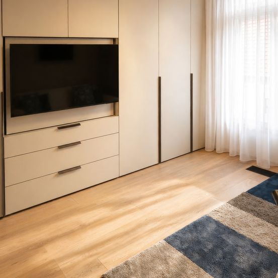 Ein modernes Wohnzimmer mit einem Flachbildfernseher, der in einem weißen Schrank montiert ist, zwei weißen Schubladen und einem weißen Vorhang. Ein Teppich liegt auf dem Holzboden.