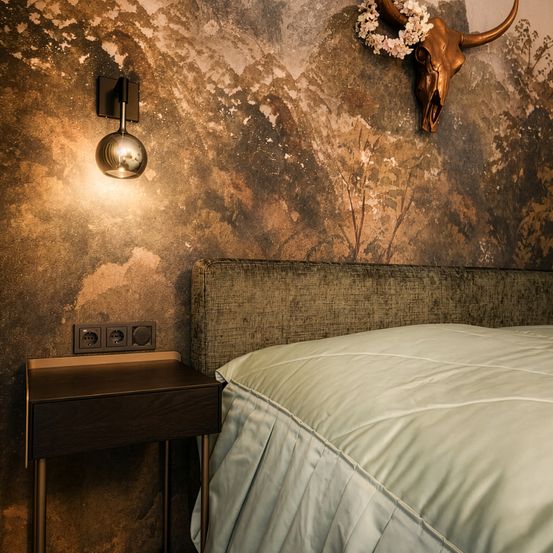 Ein Schlafzimmer mit rustikalem Wanddesign, einer Wandlampe, einem Bett mit weißer Decke und einem Nachttisch mit Steckdosen. Über dem Bett befindet sich ein montierter Bullenschädel mit Blumen.