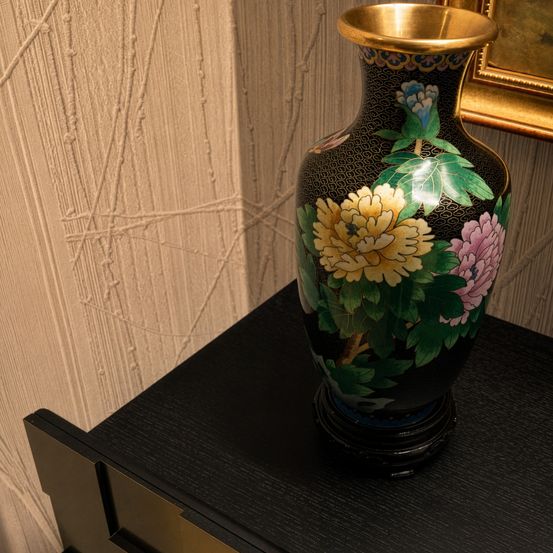 Eine schwarz-goldene Vase mit Blumenmustern steht auf einem schwarzen Tisch. Die Vase hat einen goldenen Rand und steht vor einer strukturierten Wand.