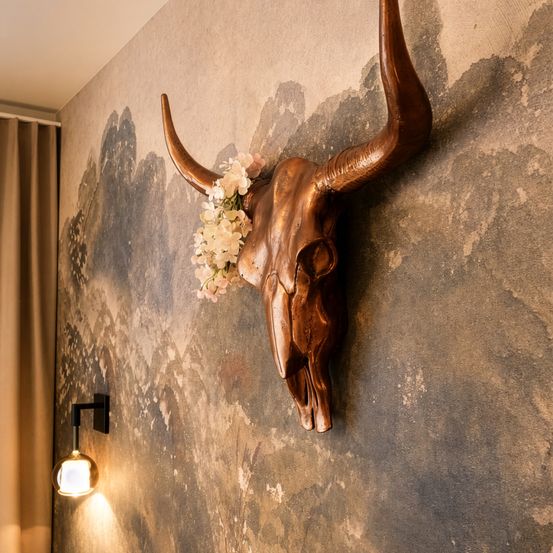 Ein kupferfarbener Bullenschädel mit Blumen ist an einer strukturierten Wand montiert. Der Schädel ist vor einem Hintergrund mit einem künstlerischen, gemalten Muster. Eine Wandlampe mit Licht ist auf der linken Seite.