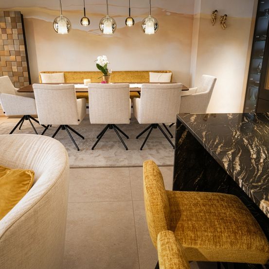 Ein Esszimmer mit beigefarbenen Stühlen um einen Tisch mit einer weißen Blume in einer Vase. Der Raum hat eine Marmorarbeitsplatte, beige Wände und eine hölzerne Wand mit einem gelben Sofa.