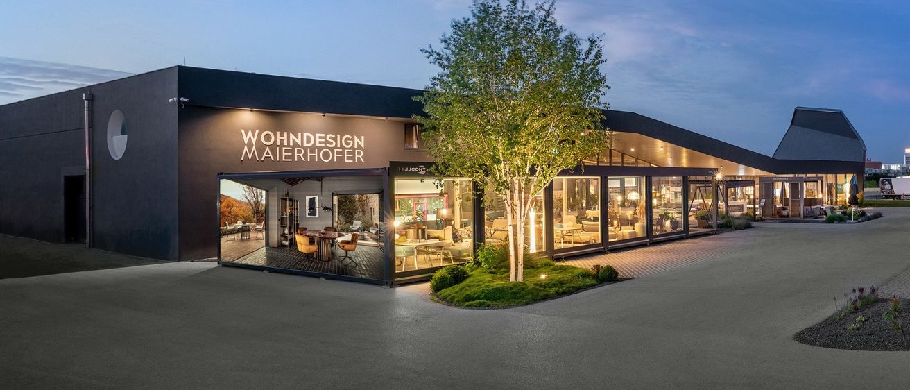 Die Vorderseite des Wohndesign Maierhofer Gebäudes ist bei Dämmerung beleuchtet und zeigt ein modernes Design mit großen Fenstern, einer Glastür und einem Baum davor.