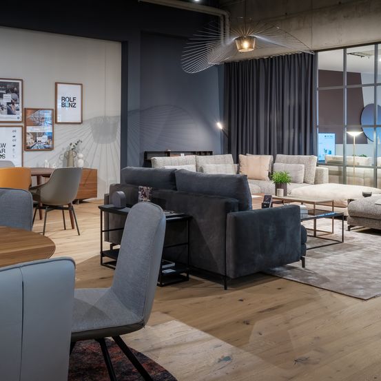Ein modernes Wohnzimmer mit einem großen grauen Sofa, einem Holztisch und gerahmten Kunstwerken an der Wand. Der Raum verfügt über einen grauen Boden und Holzmöbel.