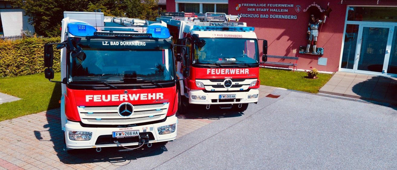 Zwei Feuerwehrfahrzeuge sind vor einem roten Gebäude geparkt, das mit 'Freiwillige Feuerwehr der Stadt Hallein Löschzug Bad Dürrnberg' beschriftet ist. Im Hintergrund sind Bäume und eine Bank zu sehen.