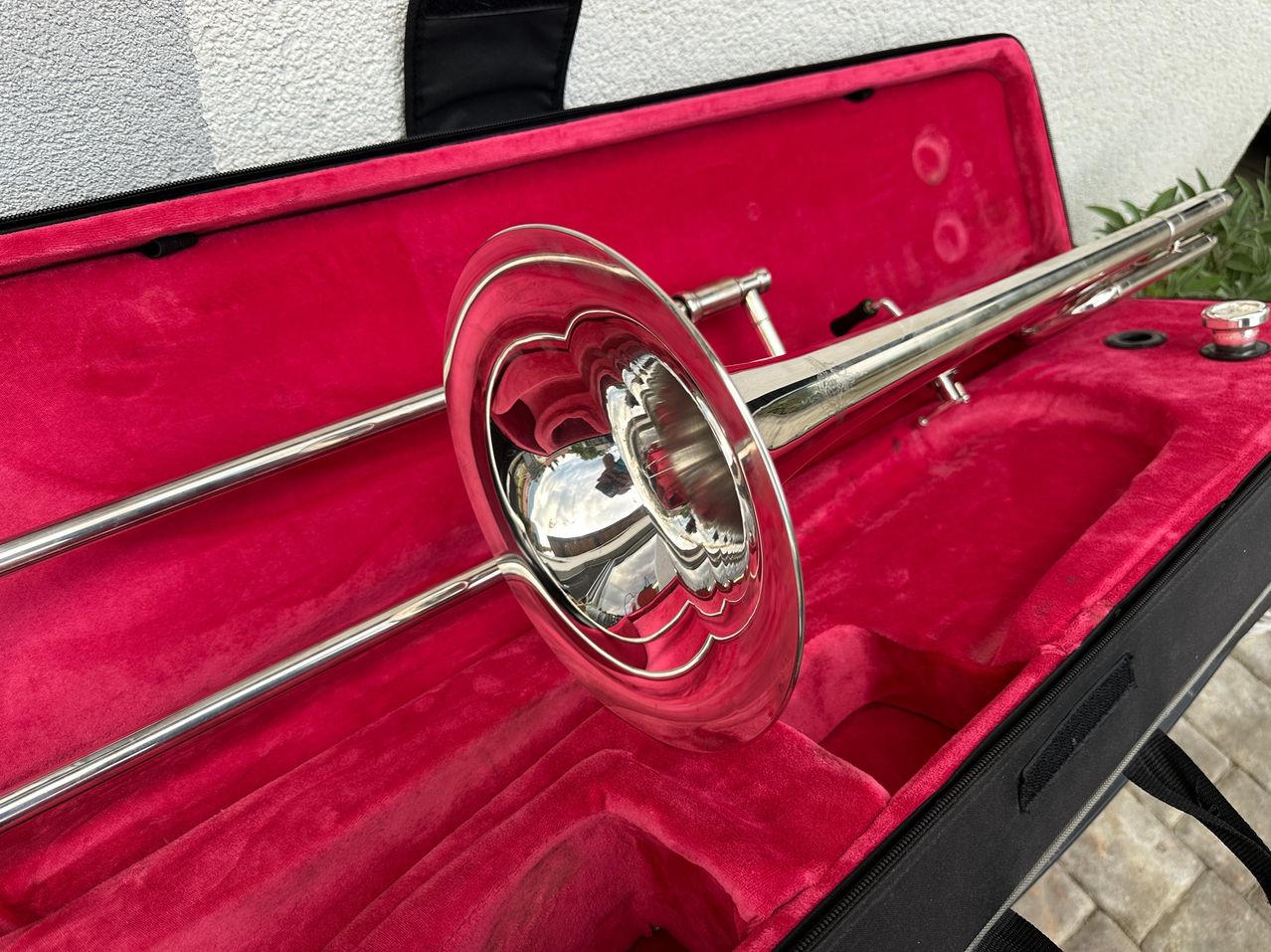 Ein polierter Posaune ruht in einem rot samtbezogenen Etui. Die glänzende Oberfläche des Instruments reflektiert das Licht, und das Etui ist mit einem schwarzen Reißverschluss verschlossen.