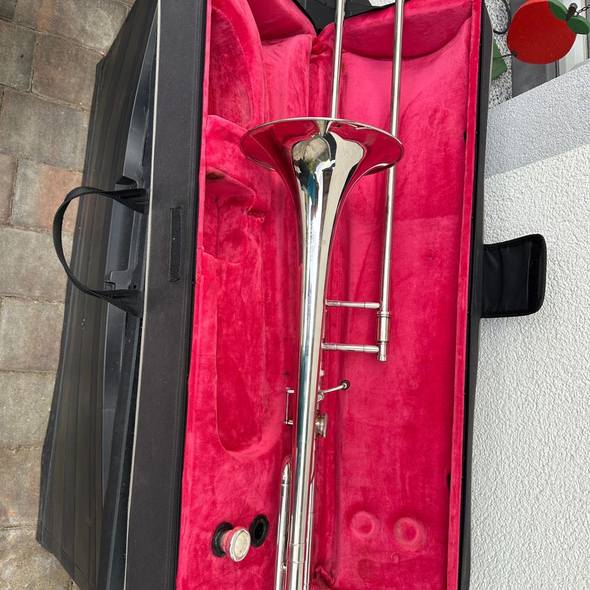 Ein Posaune befindet sich in einem schwarzen Koffer mit rotem Samt-Innenfutter. Die Posaune ist vollständig zusammengebaut und hat eine glänzende silberne Oberfläche. Der Koffer ist geöffnet und zeigt die Posaune.