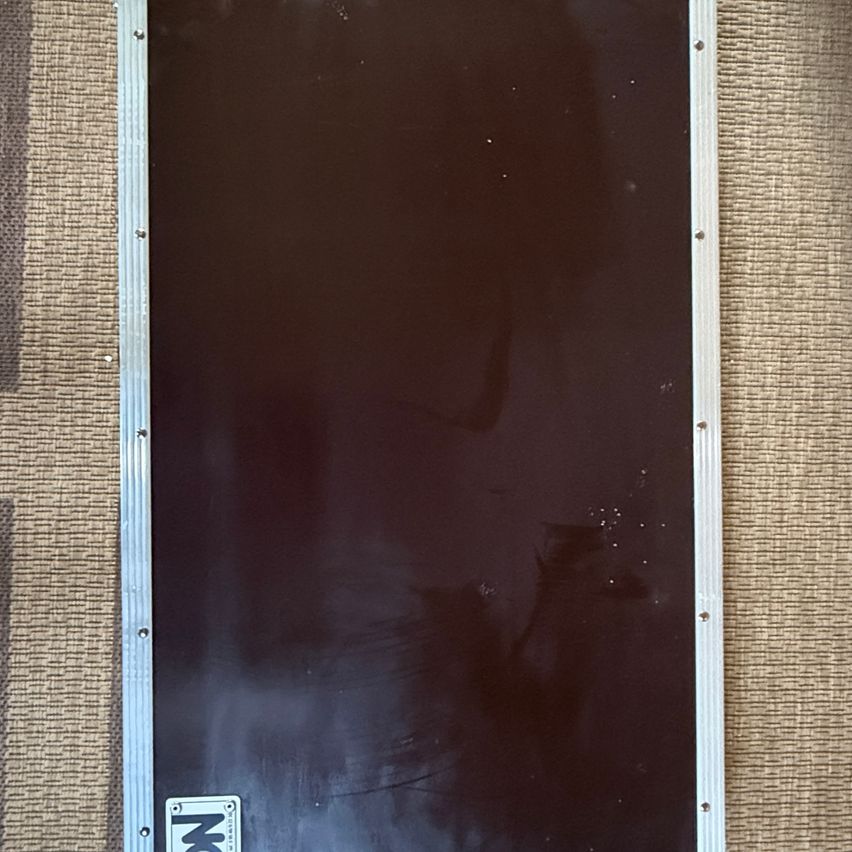 Ein dunkelfarbenes rechteckiges Objekt mit silbernen Kanten und einem kleinen weißen Aufkleber in der unteren linken Ecke steht auf einer beigefarbenen Stoffoberfläche.