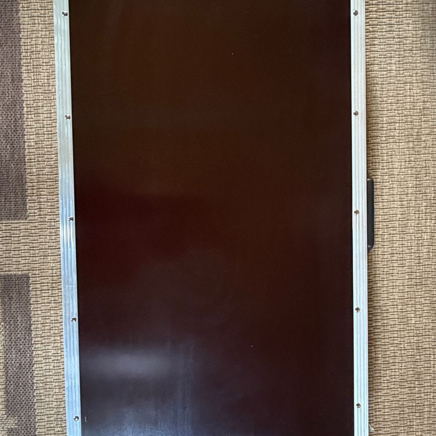 Ein rechteckiges schwarzes Objekt mit silbernen Kanten liegt auf einer beigen Oberfläche. Der Gegenstand scheint ein Monitor oder ein ähnliches Gerät zu sein, mit einer flachen Oberfläche und einem metallischen Rahmen.