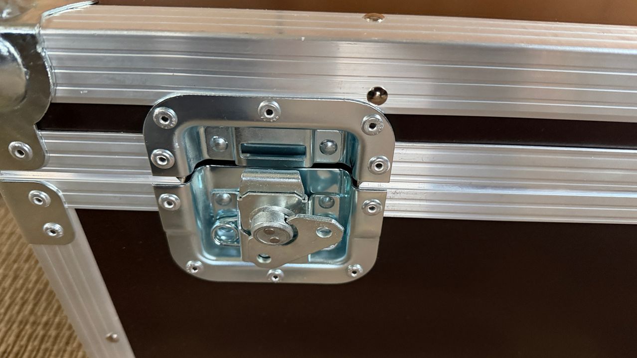 Nahaufnahme eines rechteckigen Metallkoffers mit offenem Verriegelungsmechanismus, der Schrauben und eine Scharnier zeigt.