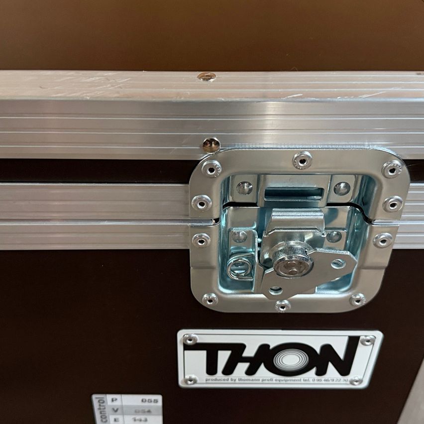 Ein Metallkoffer mit einem silbernen Verriegelungsschloss und einem weißen Etikett, das THON besagt, produziert von Thomann. Er hat einen schwarzen Rand.