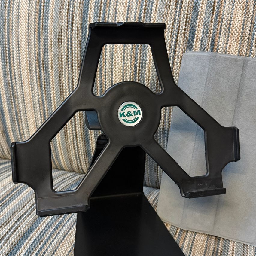 Ein schwarzer Tablet-Ständer befindet sich auf einer gestreiften Couch. Der Ständer hat ein zentrales Logo von 'K&M'.