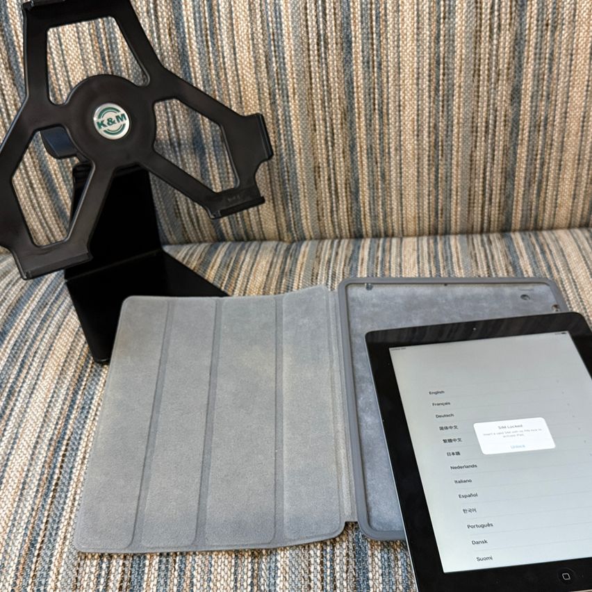 Ein Tablet liegt auf einem grauen Etui, das teilweise geöffnet ist, auf einer gestreiften Couch. Hinter dem Tablet befindet sich ein schwarzer Ständer mit einem Logo. Der Bildschirm des Tablets zeigt ein Sprachauswahlmenü an.