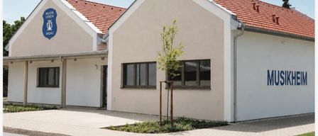 Ein Gebäude mit einem roten Dach und dem Wort Musikheim an der Seite. Es hat ein Logo auf der linken Seite. Der Text darunter lautet Einladung zur Eröffnung des neuen Musikheims am 15. August 2026, um 16 Uhr.