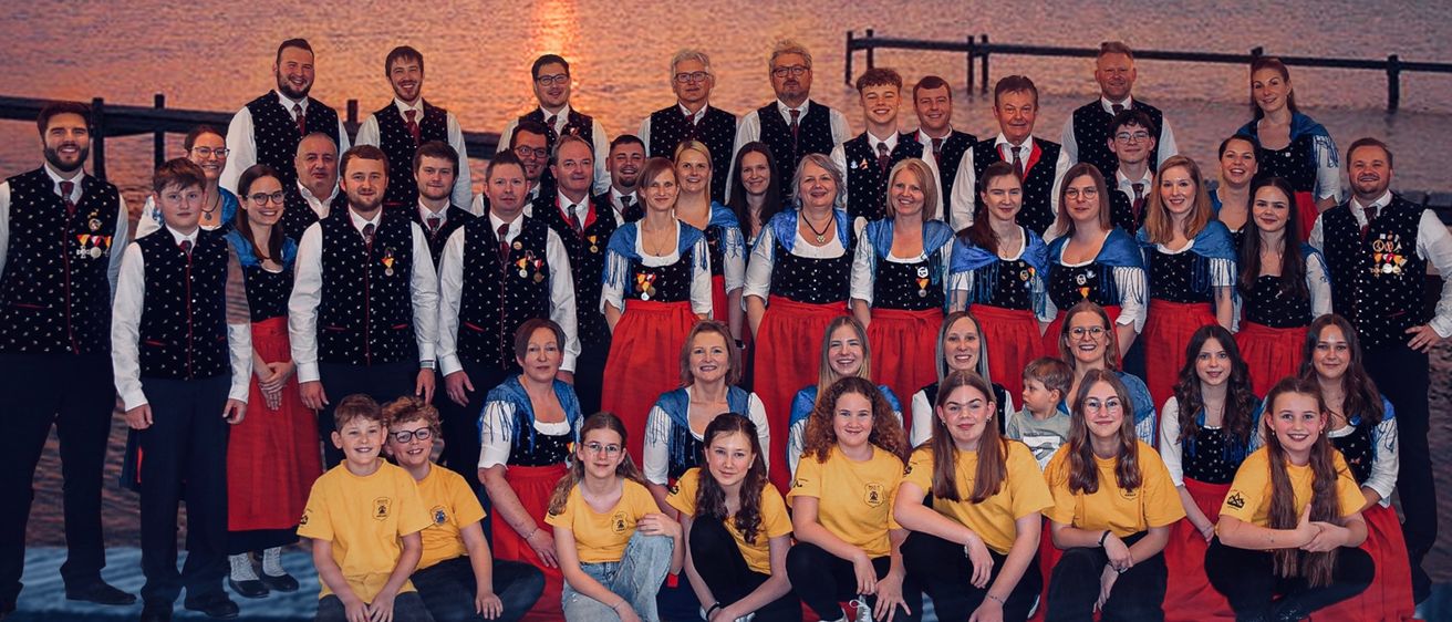 Eine Gruppe von Menschen in traditionellen Kostümen steht am Strand bei Sonnenuntergang. Sie posieren für ein Foto mit einer ruhigen See im Hintergrund.