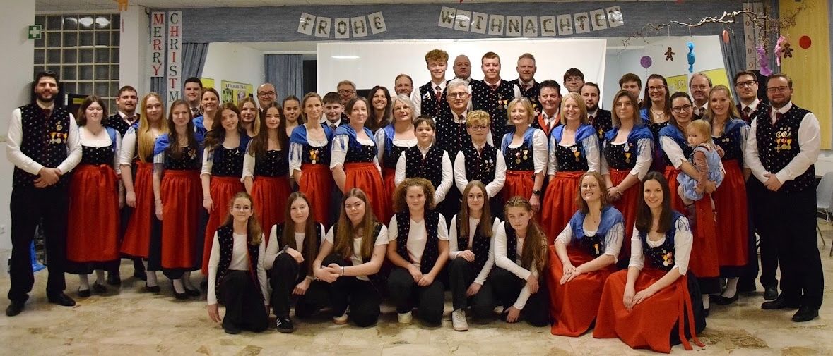 Eine Gruppe von Menschen in traditionellen Kostümen posiert für ein Weihnachtsfoto, einige stehend und andere kniend, vor einem Hintergrund mit festlichen Dekorationen.