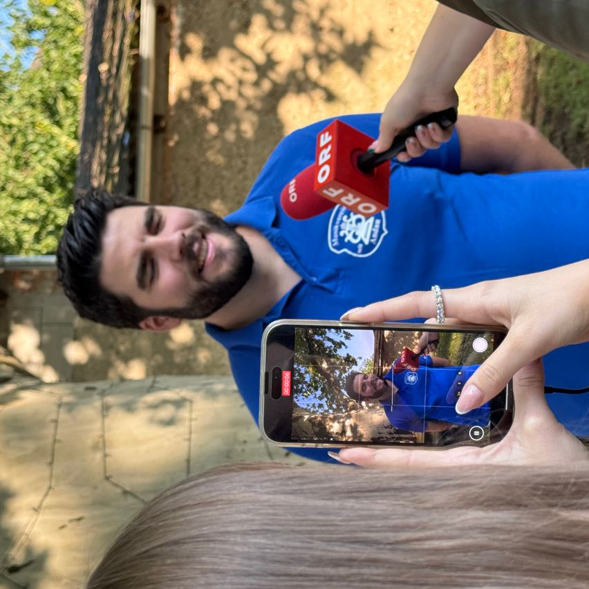 Ein Mann mit einem Mikrofon in einem blauen Shirt wird von einer Frau interviewt, die ein Telefon mit einem Selfie auf dem Bildschirm hält.