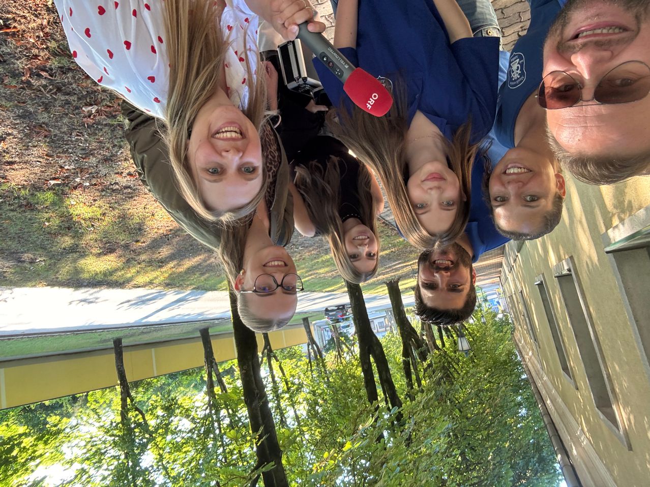 Eine Gruppe von fünf Personen macht ein Selfie im Freien. Sie lächeln und einige halten ein Mikrofon. Die Frau auf der linken Seite trägt eine Brille. Hinter ihnen sind Bäume und ein Weg.