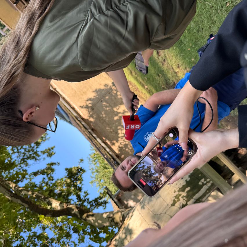Eine Person mit Brille hält ein Smartphone und zeigt ein Foto eines Mannes in einem blauen Shirt. Dahinter hält ein Mann in einem blauen Shirt ein Mikrofon. Sie befinden sich auf einem Rasenfeld mit einem Baum im Hintergrund.