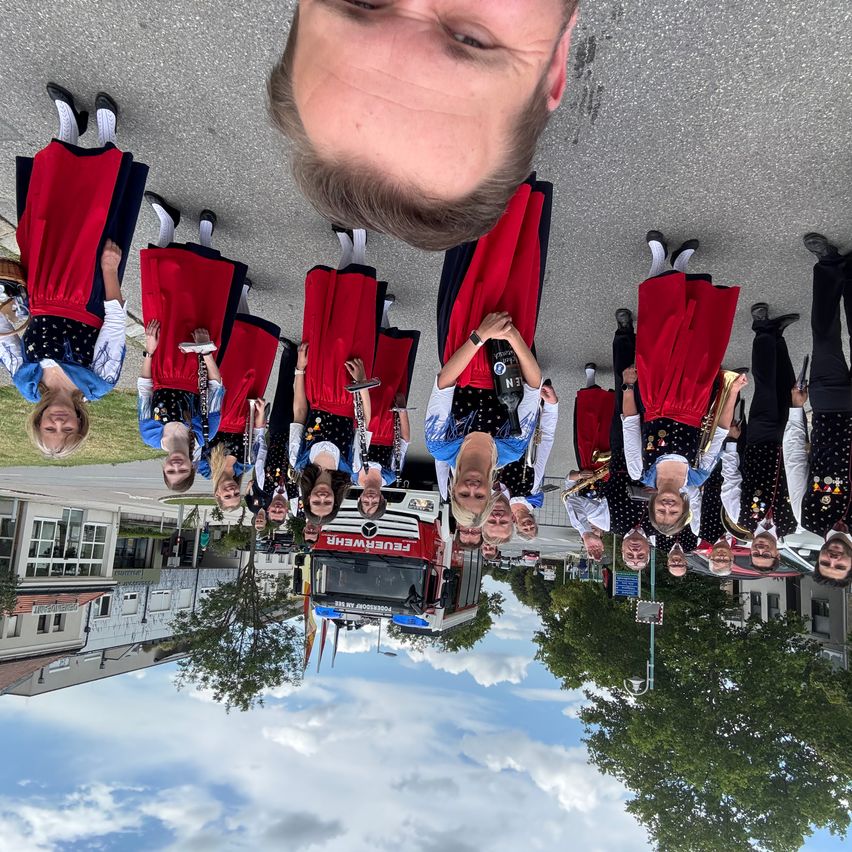 Eine Gruppe von Menschen in traditioneller Kleidung steht unter einem Selfie eines Mannes, mit einem roten LKW in der Mitte. Gebäude und Bäume spiegeln sich auf dem Boden.