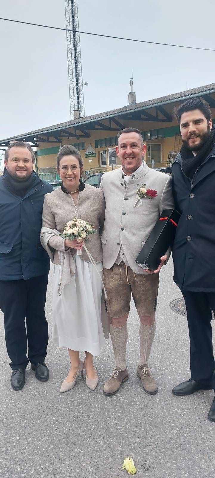 Vier Personen stehen in einer Reihe draußen. Eine Frau in einem weißen Kleid hält einen Blumenstrauß. Der Mann neben ihr trägt einen grauen Anzug und Lederhosen. Der Mann auf der rechten Seite hält eine schwarze Box.
