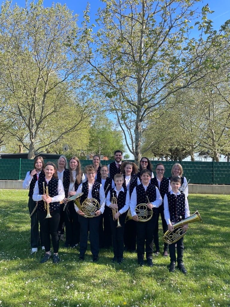 Eine Gruppe junger Musiker in gleichen Uniformen posiert mit ihren Instrumenten in einem Park, mit Bäumen und einem grünen Zaun im Hintergrund.